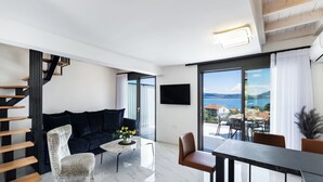 En moderne stue med en mørkeblå sofa, en hvid stol med mønster, et lille rundt bord med en vase blomster og et spiseområde med bord og stole. En trappe med trætrin er synlig til venstre, og et stort vindue giver udsigt over havet og en by i det fjerne.