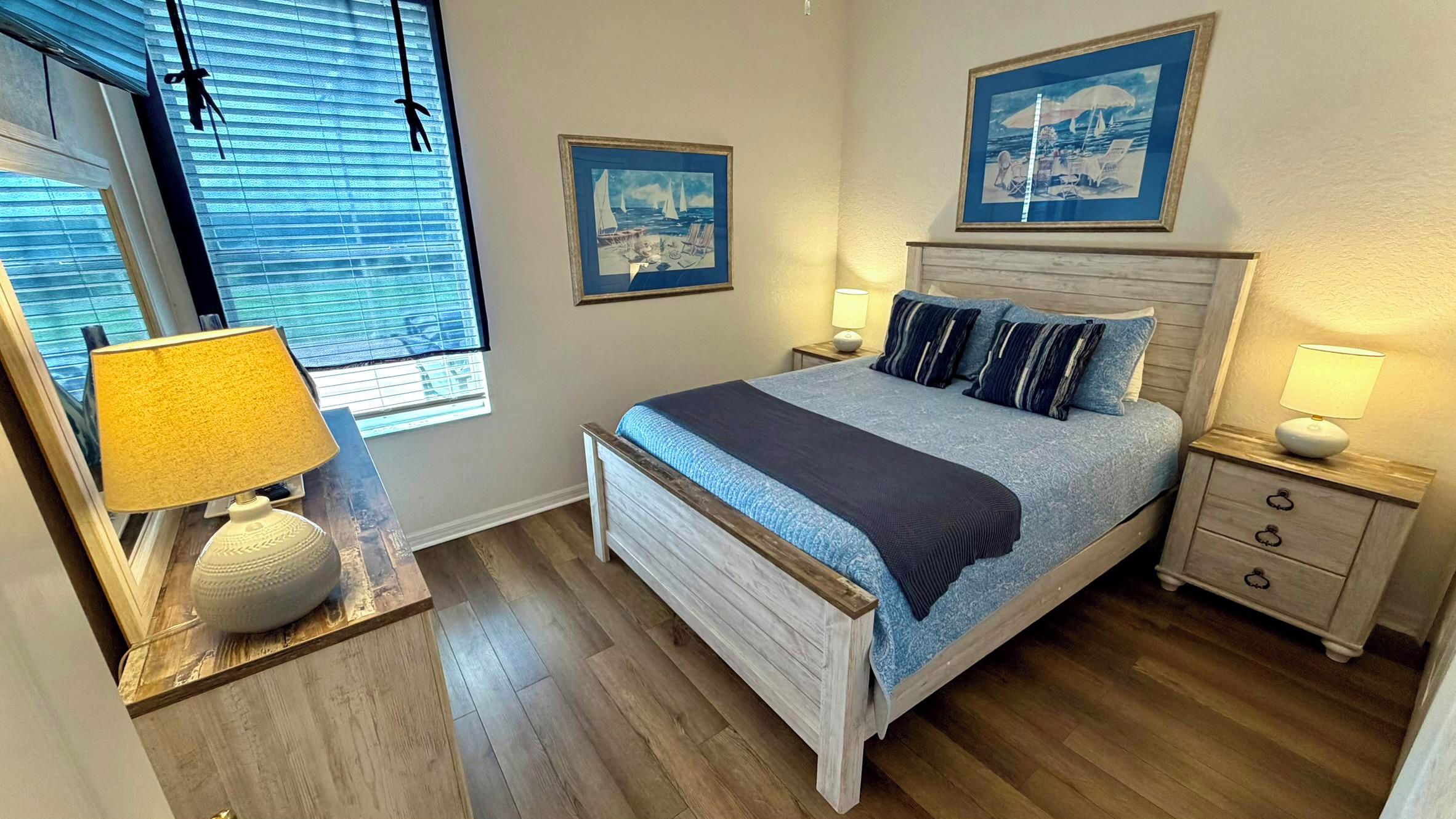 A bedroom with a bed, two bedside tables, a window with blinds, and two wall-mounted pictures.