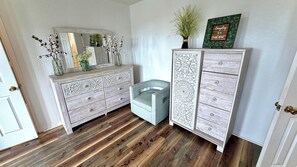 A room with a wooden floor, a white door, a mirror, a dresser with drawers, and a chair.