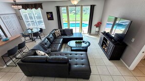 A modern living room with a sectional sofa, a coffee table, a flat-screen TV, and a dining area with a table and chairs. - Highlands Reserve 6 Bed Pool Home - Spacious, Modern & Clean (Davenport)