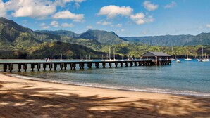 A beach with a wooden pier extending into calm waters, surrounded by lush green mountains. - Penthouse with Panoramic Ocean Views From Private Lanais (Koloa)