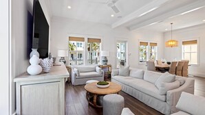 A spacious living room with a large flat-screen TV, two sofas, a round coffee table, and a dining area with a table and chairs. - Beachtown - STEPS FROM BEACH & POOL - Breathtaking 5-bedroom house w/Elevator! (Galveston)