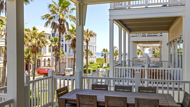 A covered outdoor seating area with wooden tables and chairs, white railings, and palm trees in the background. - Beachtown - STEPS FROM BEACH & POOL - Breathtaking 5-bedroom house w/Elevator! (Galveston)