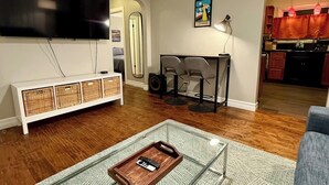 En stue med et fladskærms-TV, et glasbord, en sofa, og et køkkenområde, der kan ses igennem en åben dør.