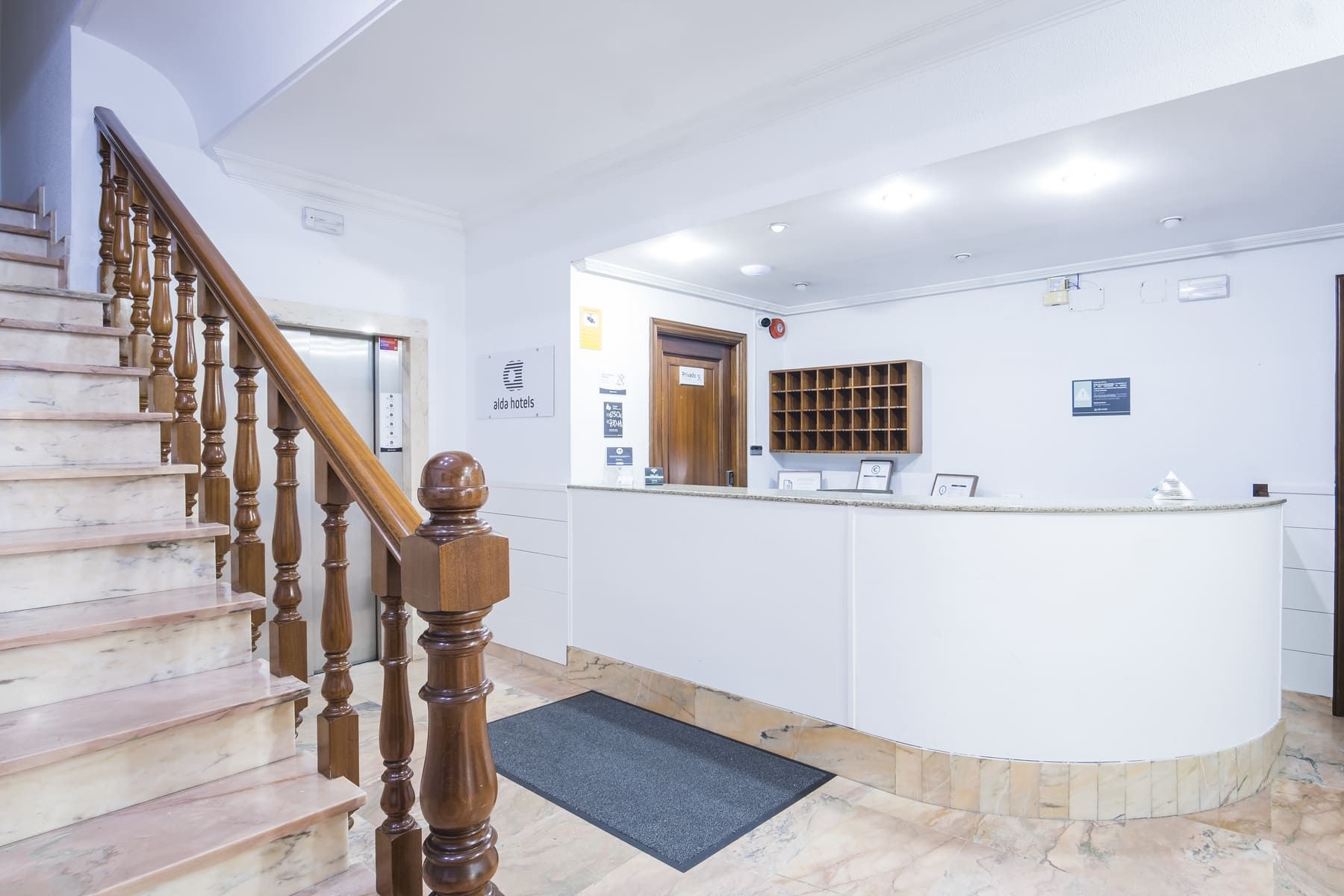 Uma área de recepção de hotel com uma escadaria de mármore, um balcão de atendimento e uma porta de madeira.