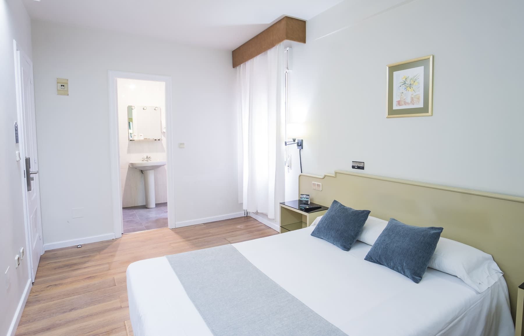 Ein Hotelzimmer mit Bett, Nachttisch, Vorhängen am Fenster, einem durchgegangenen Tür sichtbaren Badezimmer und einem gerahmten Bild an der Wand.