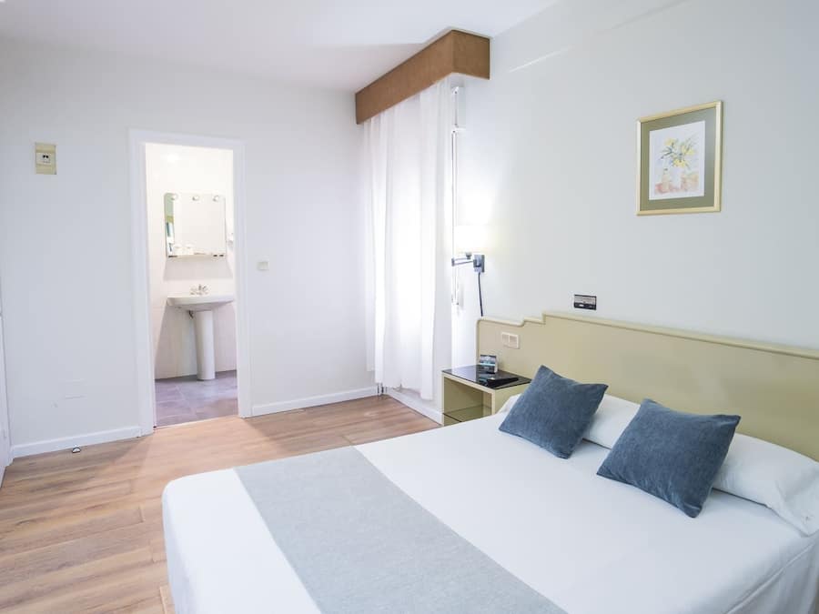 Habitación de hotel con cama, mesita de noche, cortinas en la ventana, baño visible a través de una puerta abierta y un cuadro en la pared.