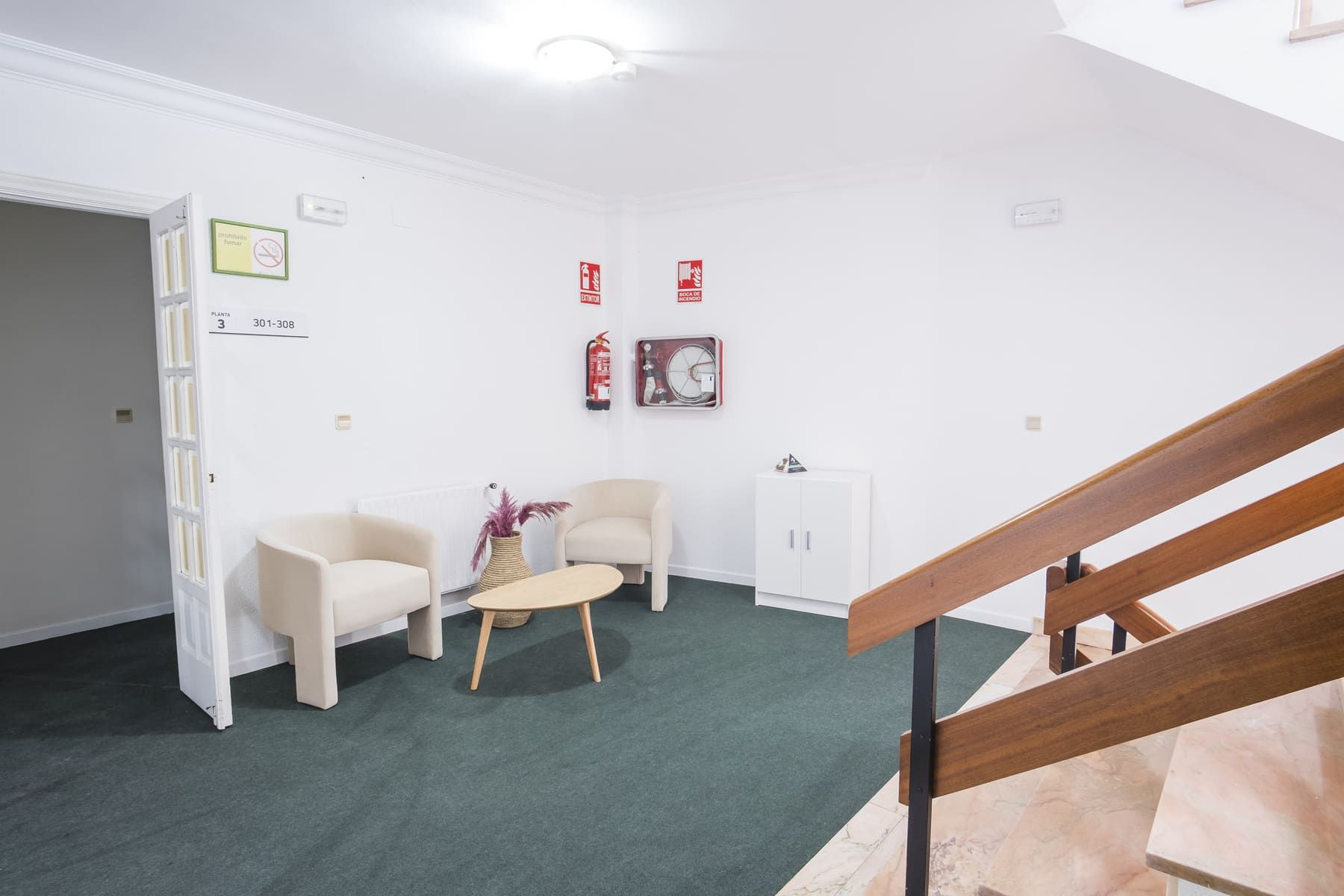 Ein Zimmer mit grünem Teppich, zwei weißen Sesseln, einem kleinen runden Tisch, einem weißen Schrank und einer Holztreppe.
