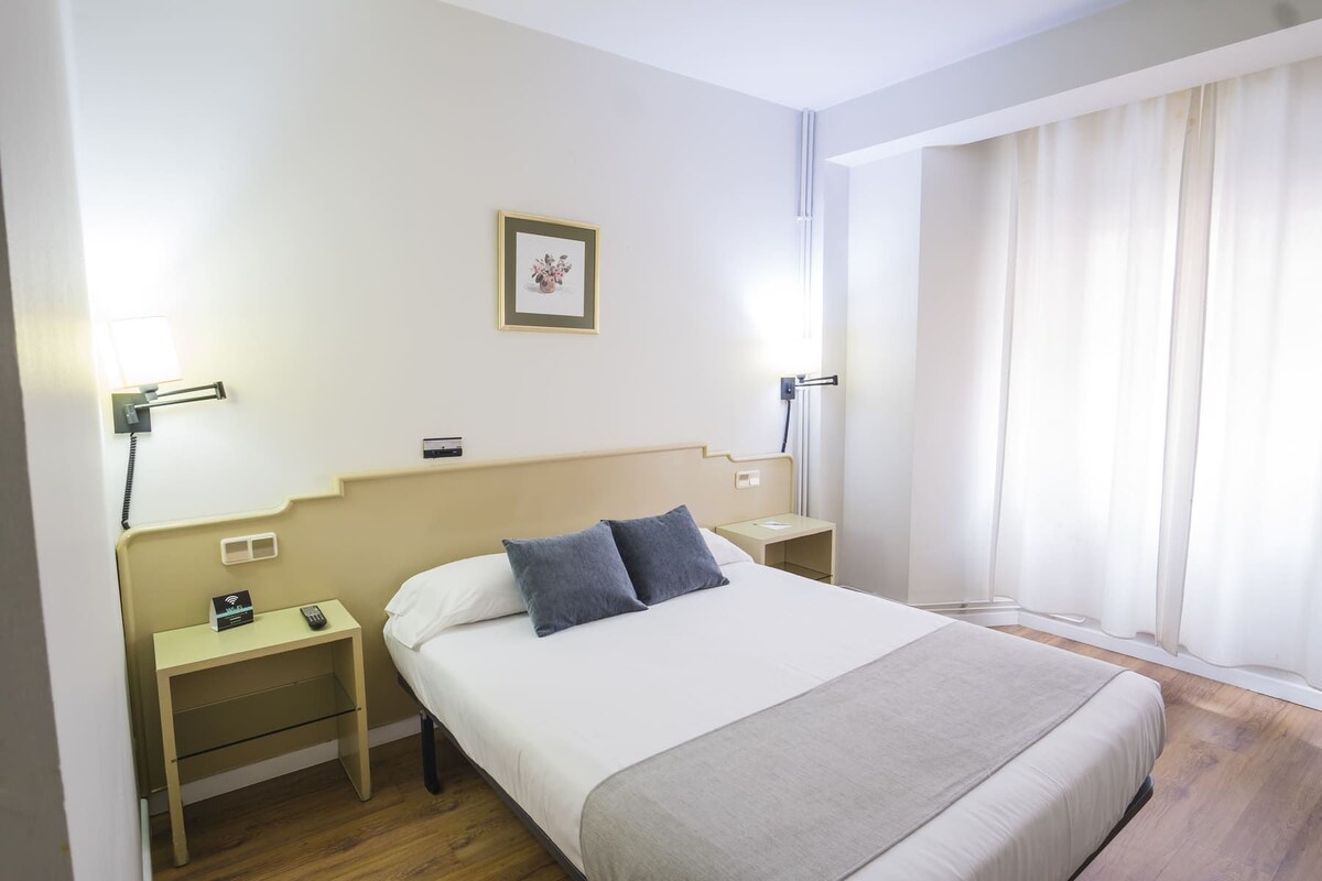 Een hotelkamer met een bed, nachtkastjes, een telefoon aan de muur, een schilderij en een raam met gordijnen.