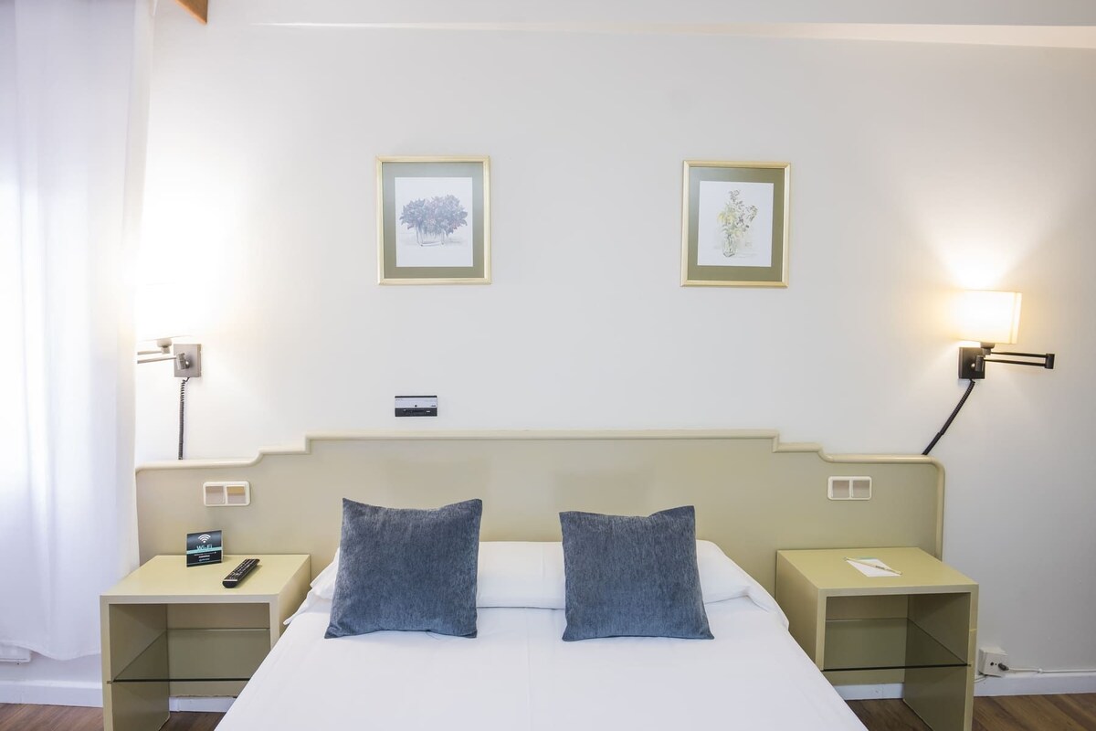 A hotel room with a bed, two pillows, a nightstand with a phone, a bedside table with a lamp, and two framed pictures on the wall.