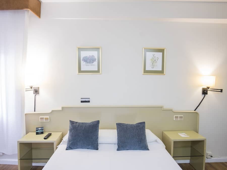Habitación de hotel con cama, dos almohadas, mesita de noche con teléfono, mesita de noche con lámpara y dos cuadros en la pared.