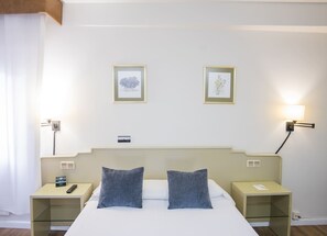 Une chambre d’hôtel avec un lit, deux oreillers, une table de chevet avec un téléphone, une autre table de chevet avec une lampe, et deux tableaux encadrés au mur.