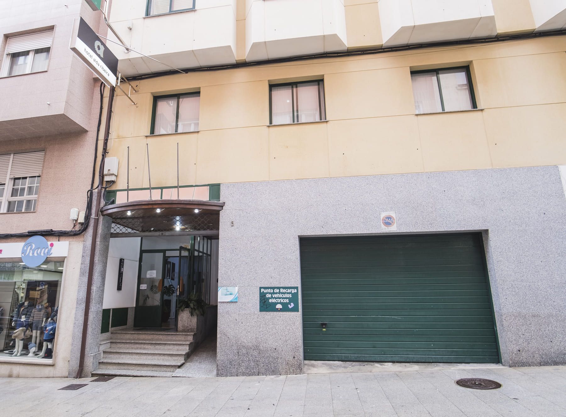 Un edificio con una puerta de garaje verde, un letrero que indica un punto de estacionamiento para vehículos eléctricos y un escaparate con maniquíes.