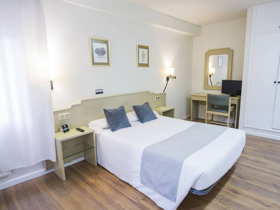 Habitación de hotel con cama, mesitas de noche, escritorio con silla, armario y dos cuadros en la pared.