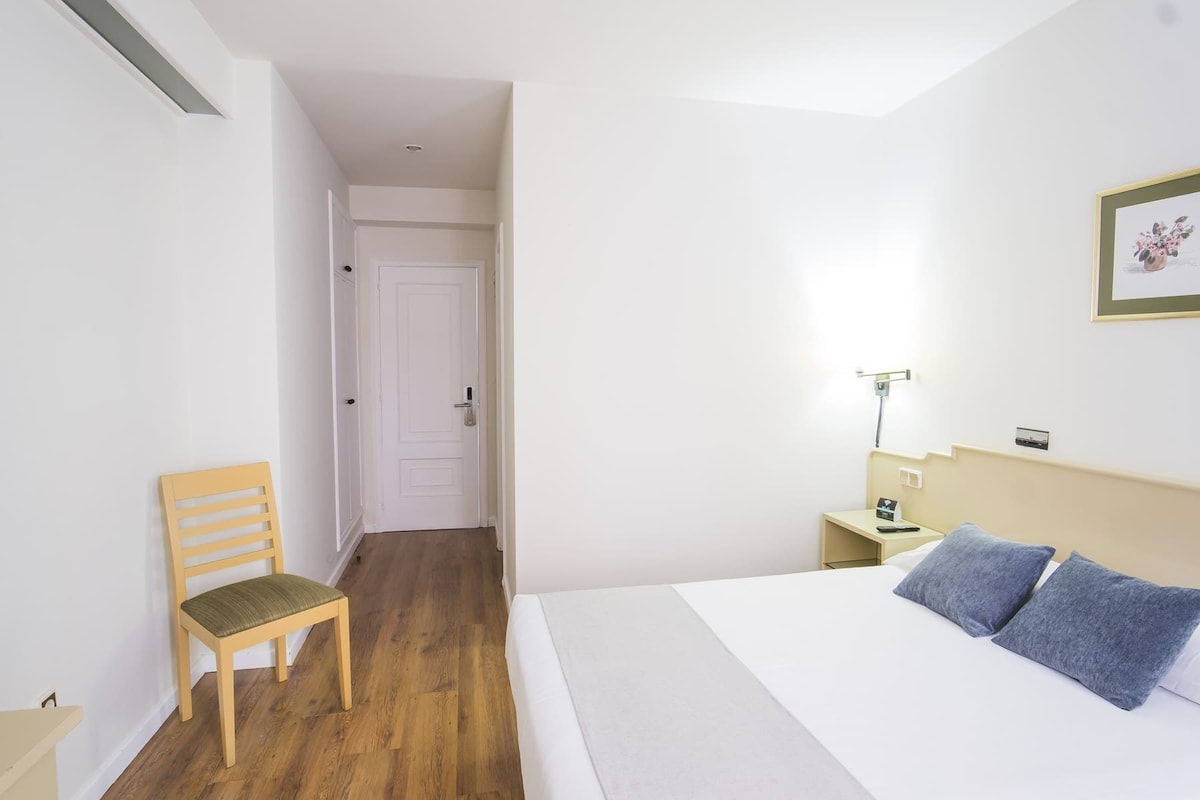 Quarto de hotel com uma cadeira de madeira, cama com almofadas azuis, mesa de cabeceira com abajur e porta que dá para outro quarto.