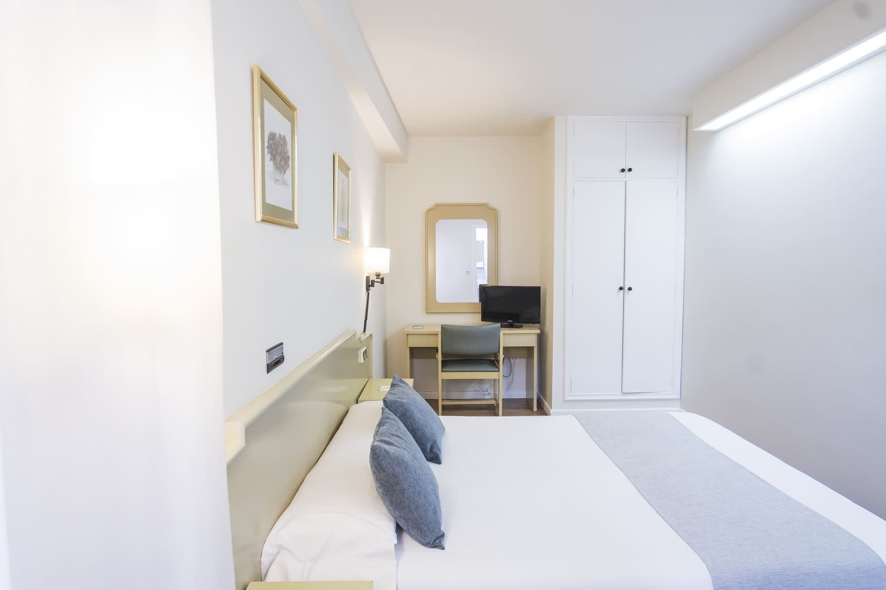 A hotel room with a bed, a desk with a computer, a chair, a wardrobe, and a mirror on the wall.