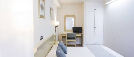 A hotel room with a bed, a desk with a computer, a chair, a wardrobe, and a mirror on the wall.