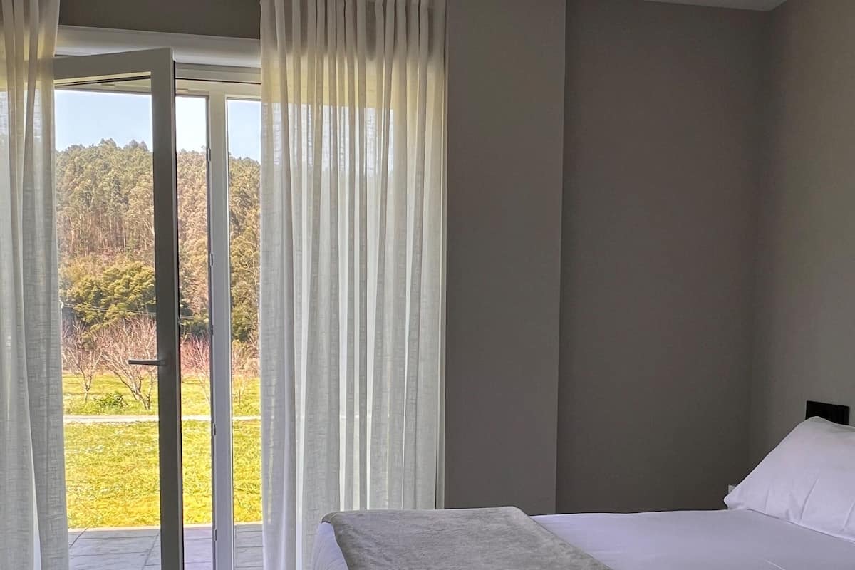Ένα υπνοδωμάτιο με ένα κρεβάτι, θέα σε ένα καταπράσινο τοπίο μέσα από μια γυάλινη πόρτα και ένα φωτιστικό οροφής.