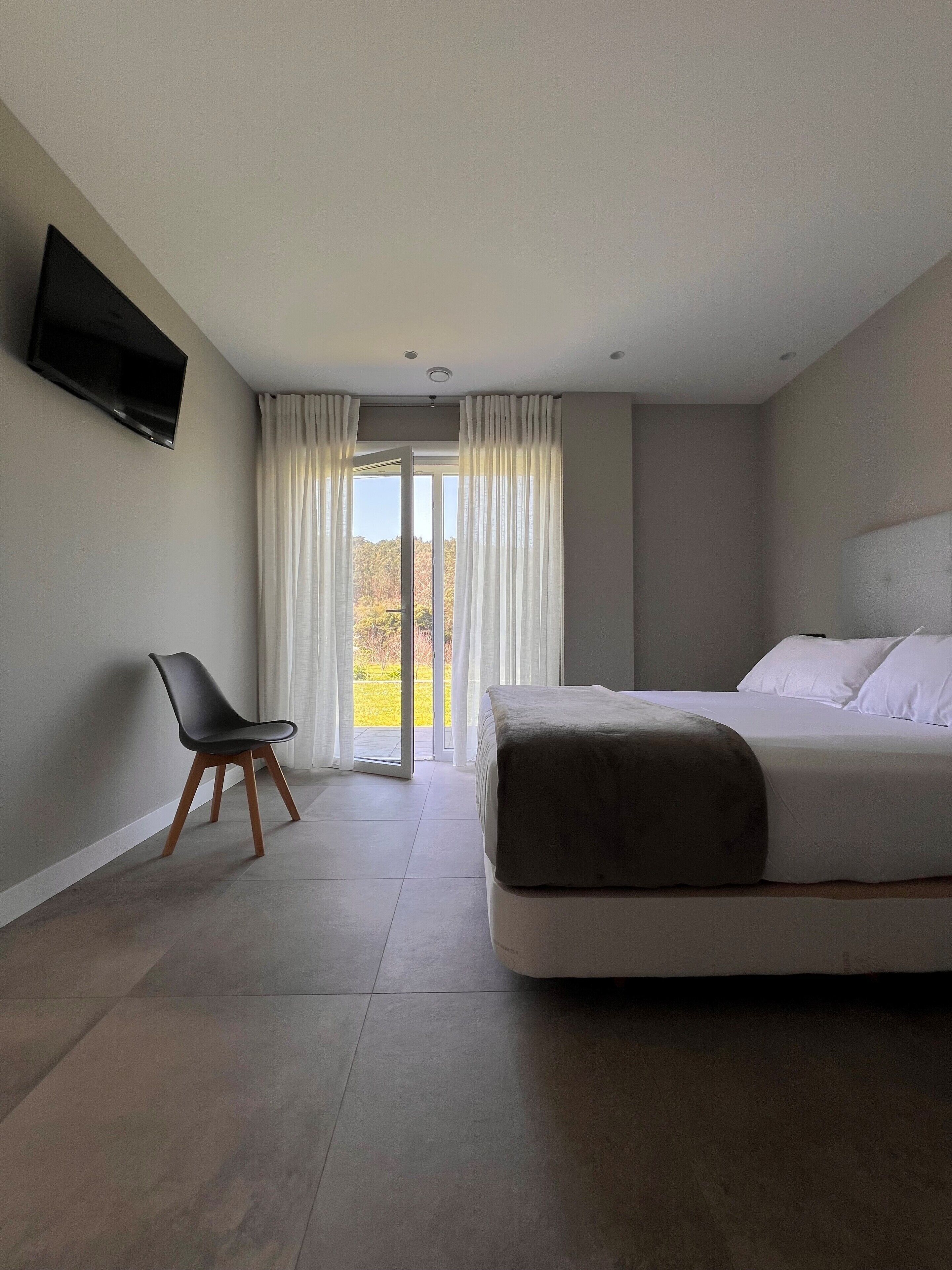 Una habitación de hotel moderna con una cama grande, un televisor de pantalla plana montado en la pared, una silla y un ventanal corredero que da a un balcón.