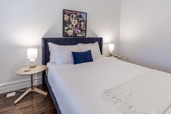 Ein ordentlich bezogenes Bett mit weißer und blauer Bettwäsche, zwei Nachttische mit Lampen, ein gerahmtes Blumenbild über dem Bett und ein Holzboden.