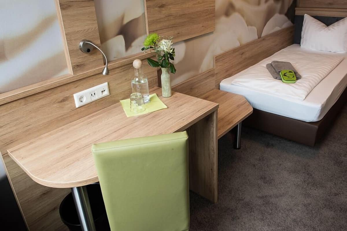 A hotel room with a wooden desk, a green chair, a bed with a white mattress, and a small bedside table with a green object.