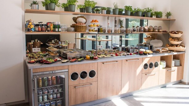 A kitchen with wooden cabinets, a built-in refrigerator, and shelves stocked with food items and plants. - The Outlet Hotel (Metzingen)