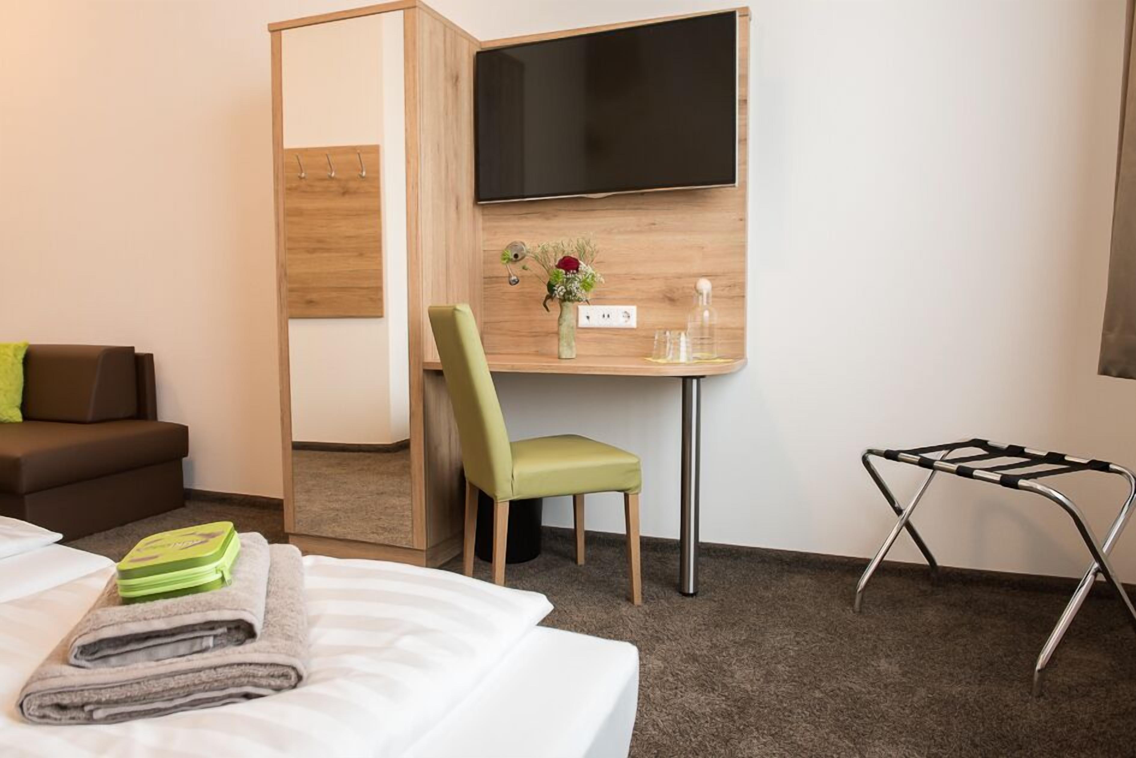 Habitació d'hotel amb llit, escriptori amb cadira, televisió penjada a la paret i taula petita amb un vas de flors.