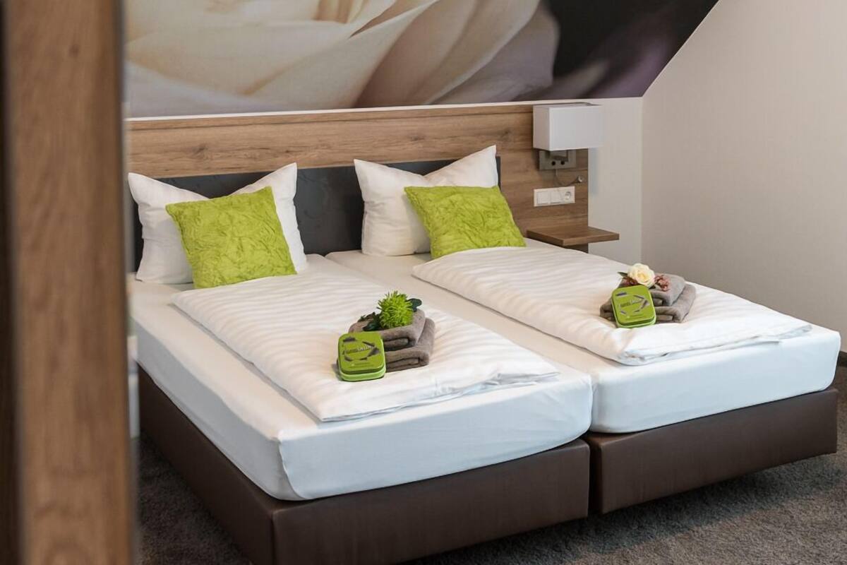 A double bed with white bedding and green pillows, a small bedside table with a lamp, and a wall-mounted outlet.