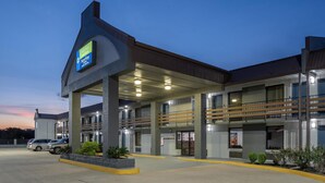 Ein Hotel mit überdachter Eingangspartie, mehreren Balkonen und einem Parkplatz mit Autos.