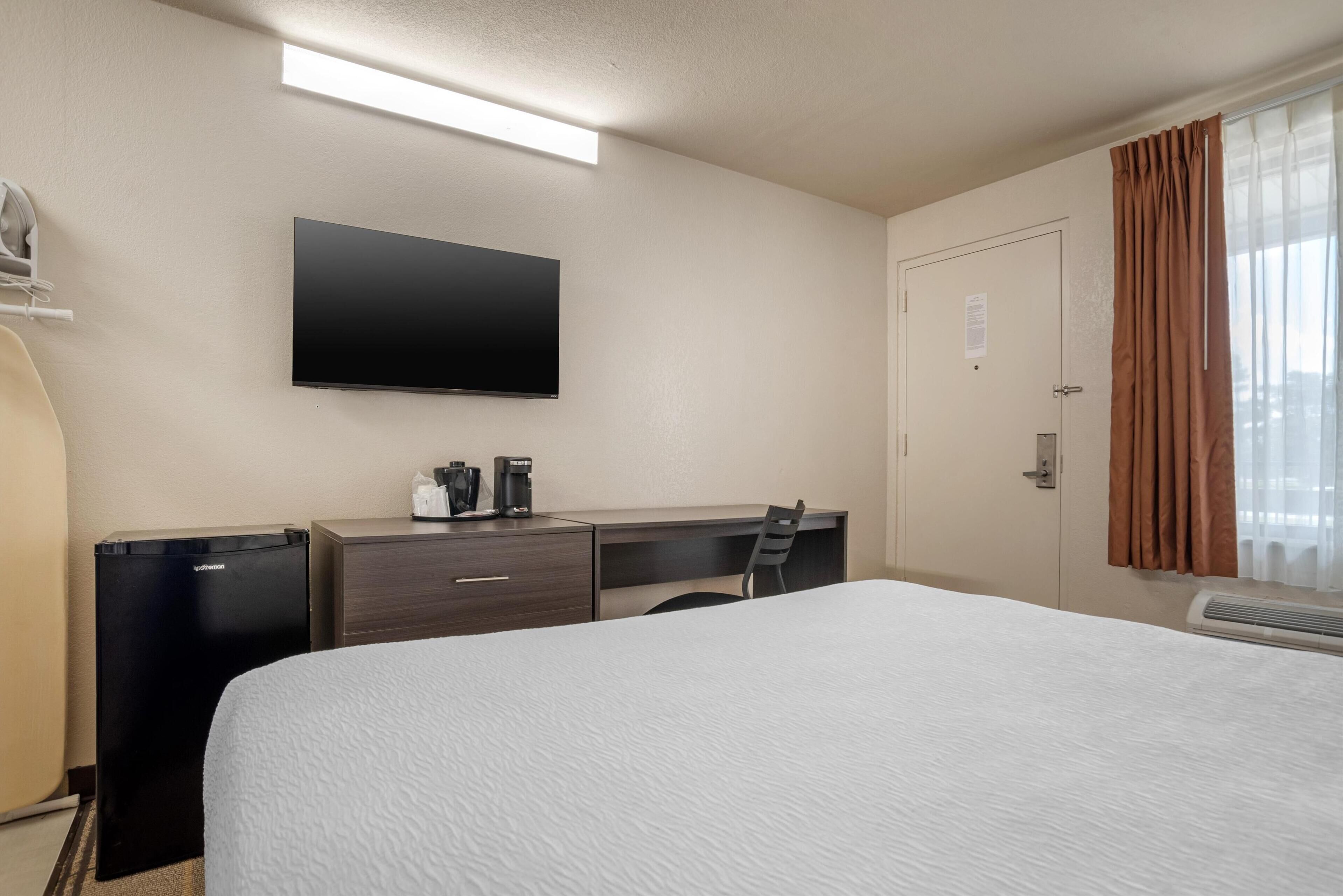 Ein Hotelzimmer mit einem Bett, einem Schreibtisch mit Stuhl, einem Flachbildfernseher, einer Minikühlschrank und einem Fenster mit Vorhängen.