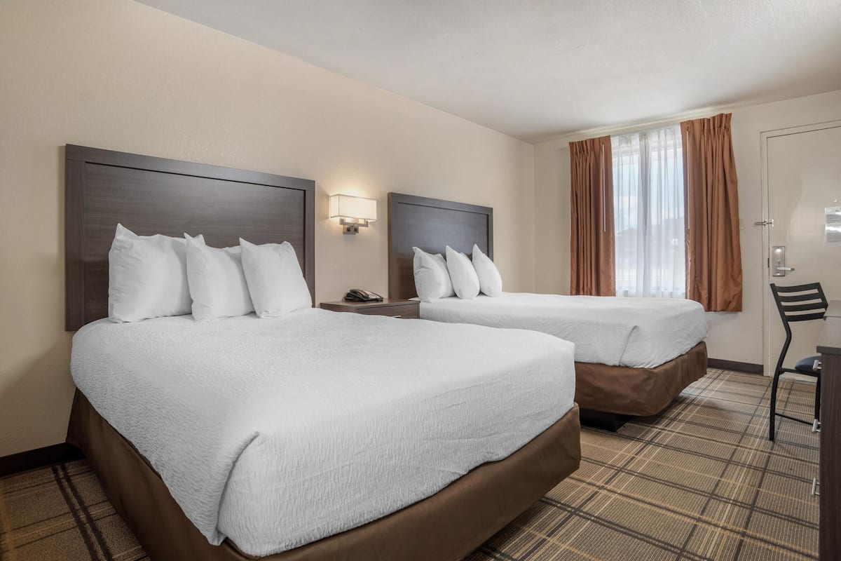 A hotel room with two beds, a nightstand, a lamp, a chair, and a window with curtains.