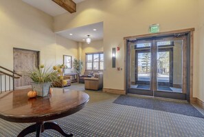 A hotel lobby with a reception desk, a large wooden table with a vase of flowers, a seating area with a sofa and armchairs, and a glass door leading outside.