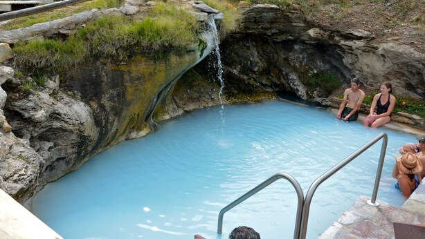 Une source d’eau chaude naturelle, avec une petite cascade, entourée d’un paysage rocheux et de verdure.