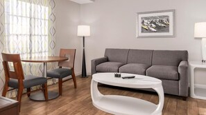 Ein modernes Wohnzimmer mit einem grauen Sofa, einem runden Couchtisch, einem Holzessttisch mit Stühlen, einer Stehlampe und einem gerahmten Bild an der Wand.