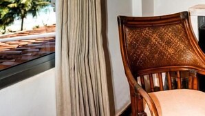Una habitación con vista a palmeras, una ventana con cortinas, una silla de madera y un equipo de aire acondicionado.