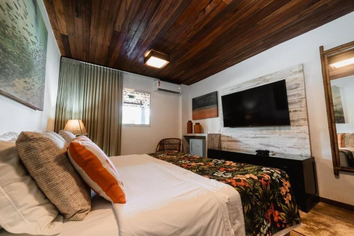 Um quarto com teto de madeira, cama com roupa de cama branca e laranja, televisão sobre um móvel de madeira e um quadro na parede.