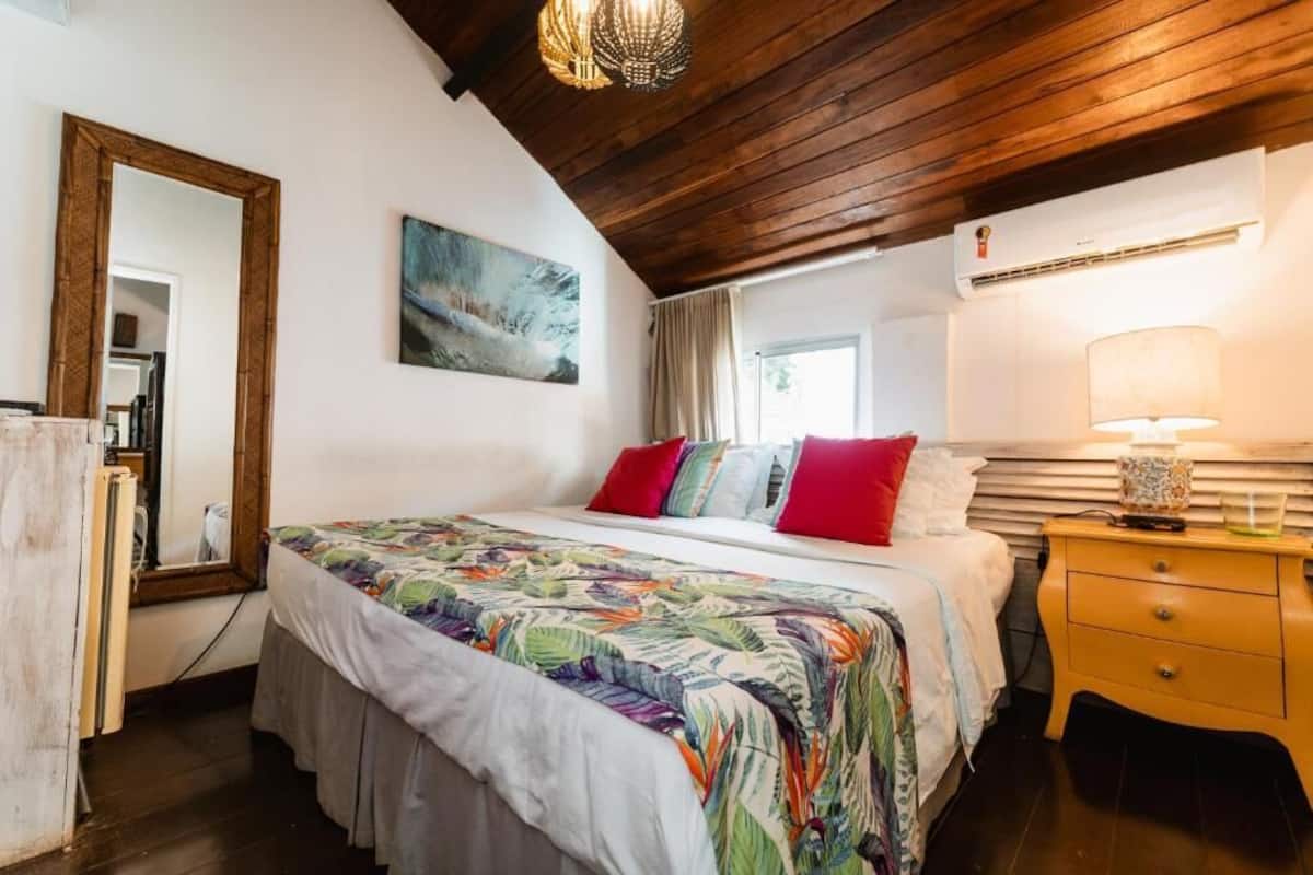 Quarto com teto de madeira, cama com roupa de cama colorida, abajur e um quadro na parede.