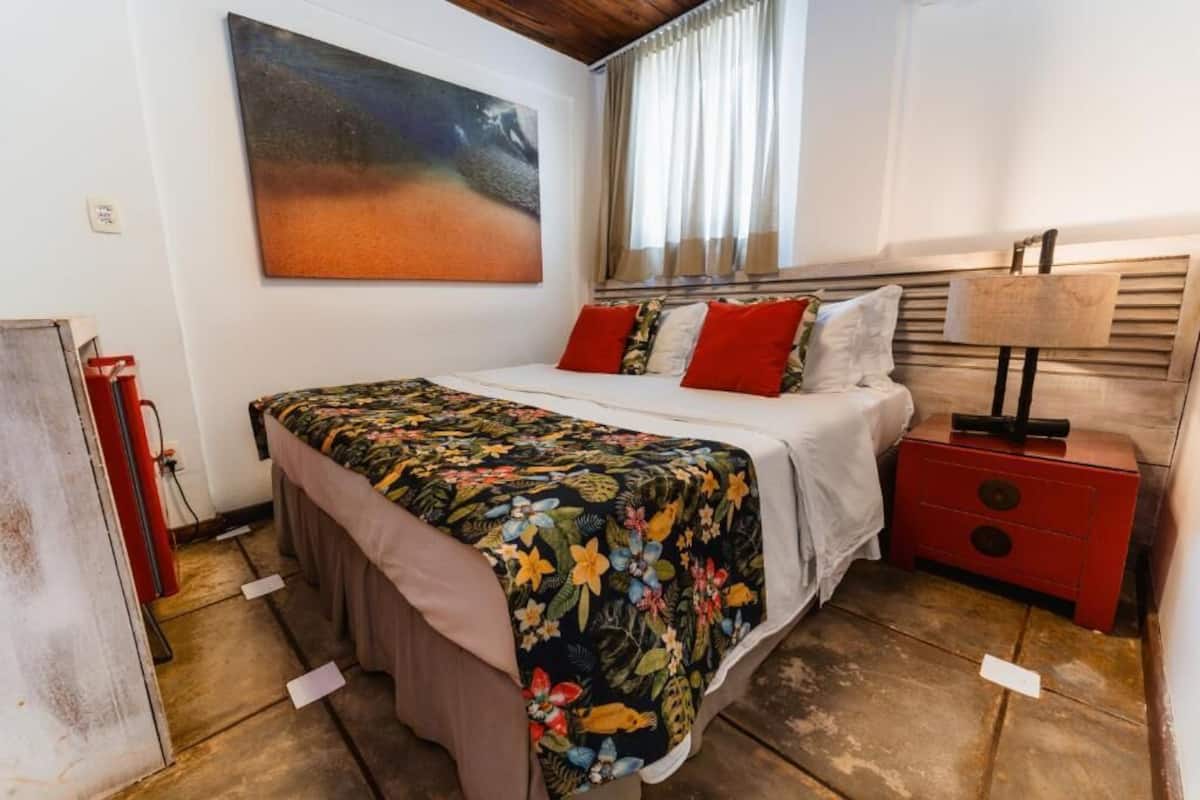 Quarto com cama, mesa de cabeceira, luminária, quadro na parede e janela com cortinas.