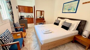 Une chambre d’hôtel avec un lit, une table de chevet en bois, une chaise, un bureau et, au fond, un coin cuisine.