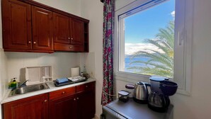 A kitchen with wooden cabinets, a sink, a window with a view of palm trees and the sea, and various kitchen appliances. - Cozy studio near the beach (Avlakia)