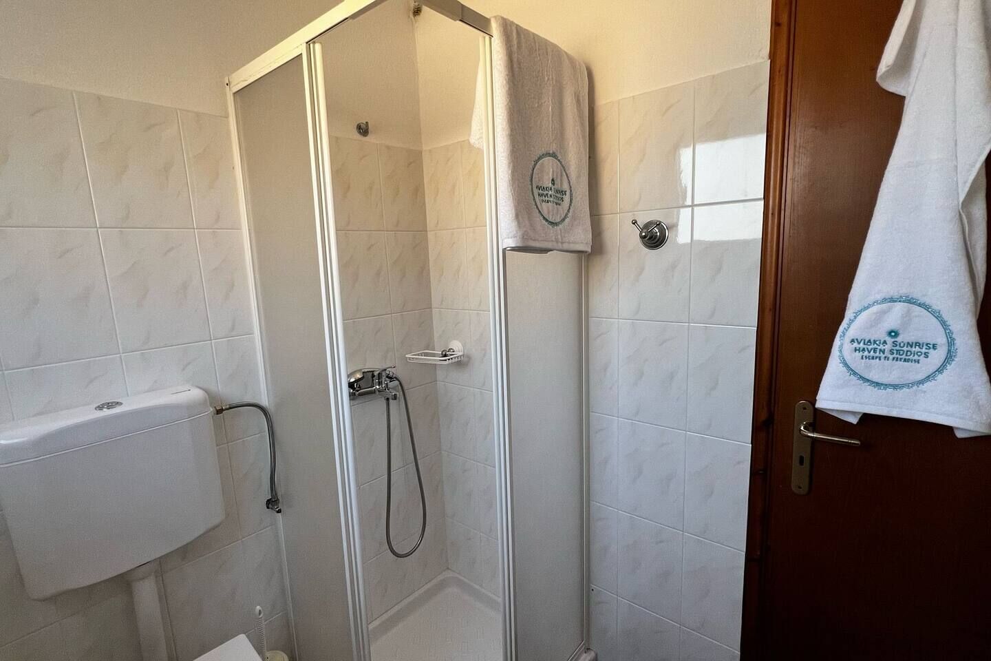 Area doccia con porta in vetro, un asciugamano bianco appeso al muro e una porta bianca con un asciugamano.
