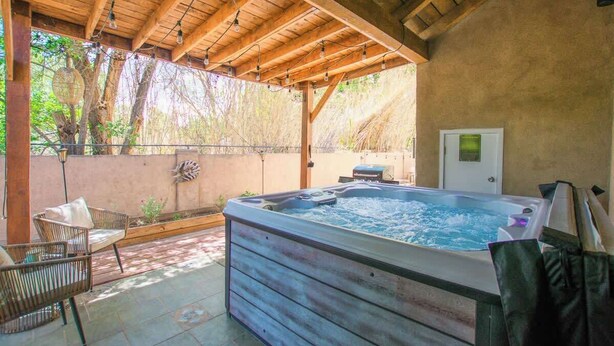Un jacuzzi bajo una pérgola de madera con sillas de mimbre, una terraza de madera y una pared con una puerta y un ventanal pequeño.