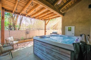 Een hottub onder een houten pergola, met rieten stoelen, een houten terras en een muur met een deur en een klein raam.