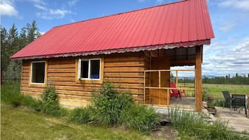 Enjoy a peaceful stay in this cozy cabin with a porch, a red chair, and a table, surrounded by a grassy field and trees.