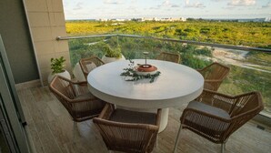 Une table ronde entourée de chaises en osier, sur un balcon doté d’une balustrade en verre, d’où l’on peut admirer un paysage verdoyant et des bâtiments éloignés.