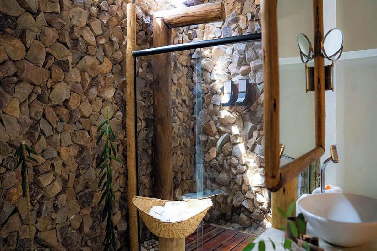 A rustic bathroom with a stone wall, a wooden shower, a hanging mirror, and a white sink.