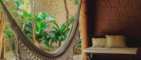 A hammock in a room with a wooden floor, a bed with pillows, and a stone wall.