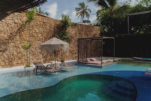 Una piscina con sillones de descanso, un muro de piedra y una zona sombreada con toldo.