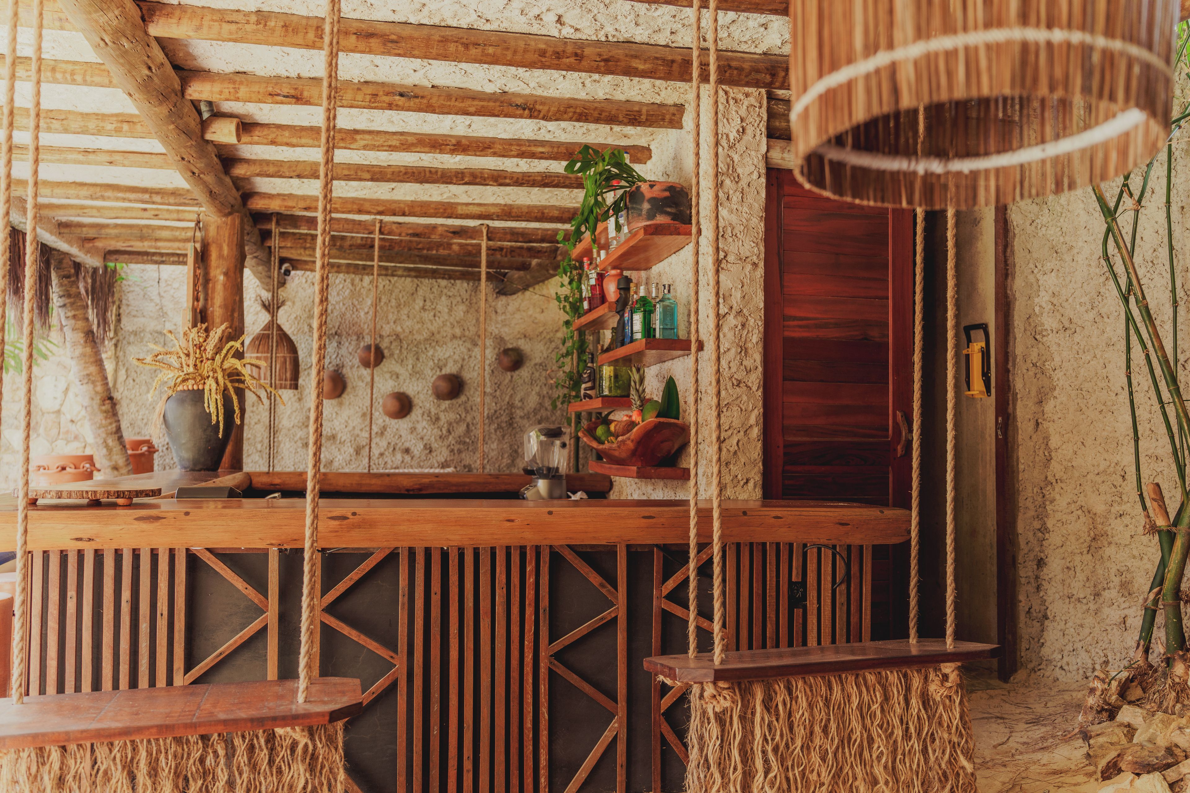 A rustic wooden bar with rope swings, a hanging light fixture, and shelves with bottles and plants.