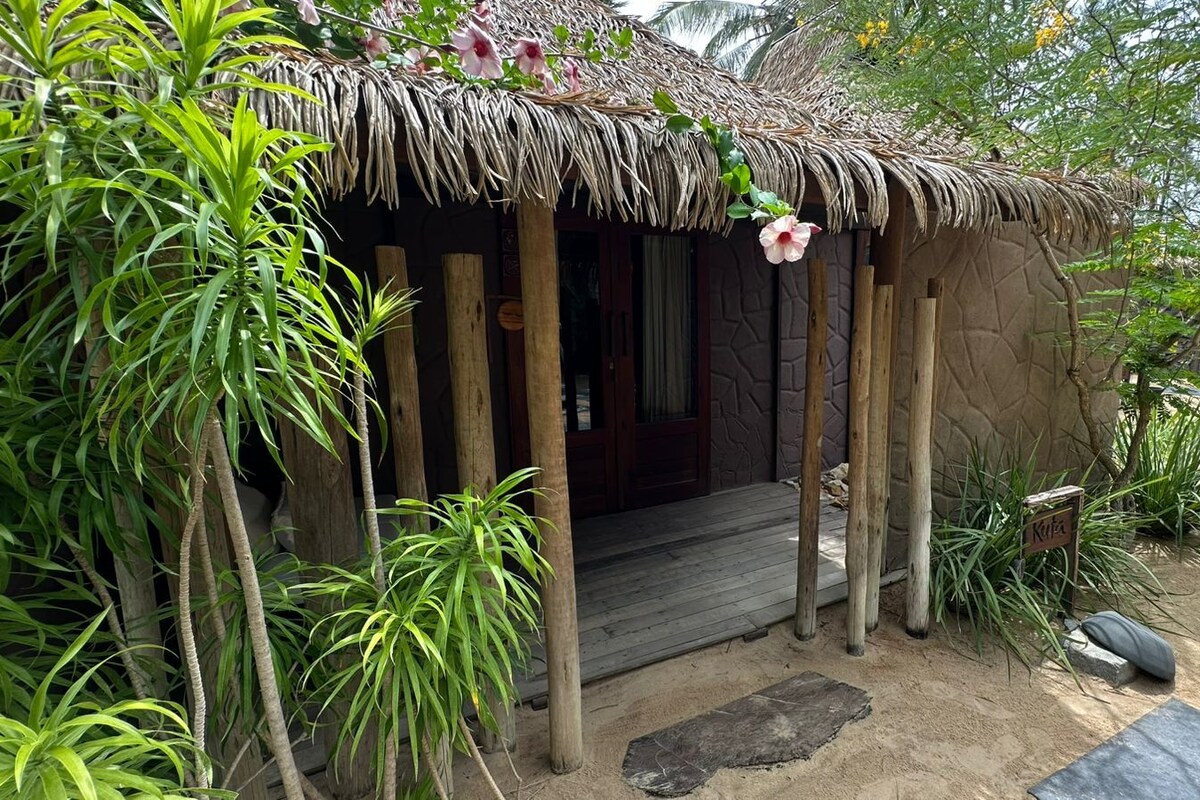 A rustic hut with a thatched roof, surrounded by tropical plants and a wooden deck.